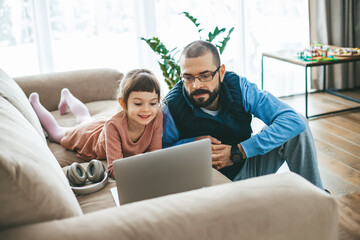 Seated side by side on a couch, a father and his daughter both engrossed in a laptop screen,...