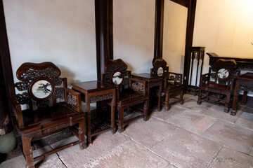 A traditional and historic Chinese living room with ornate wooden furniture. Four intricately carved chairs and small tables are arranged in a symmetrical pattern on a stone floor.