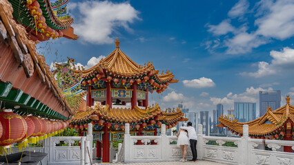 Obraz premium Wedding in Malaysia.The bride and groom are hugging each other on the terrace of a Chinese temple, looking at the city. Tiled roofs with rows of red lanterns along the curved edges, a carved fence.