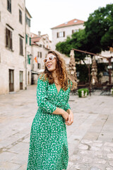 Naklejka premium A woman in a green dress enjoys a sunny day in the charming historic square of a Mediterranean town, gazing at the tall buildings and clock tower. Fashion, style, tourism concept.