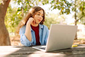 Young  freelancer woman in casual attire focused on laptop while sitting at a park table on a sunny day, enjoying a moment of productivity. E-learning concept. Remote work.