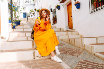 A young traveler woman in an orange dress enjoys using her smartphone while strolling through a charming whitewashed street in a sunny village. Travel, tourism, technology, blogging.