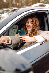 A young woman with curly hair enjoys driving by smiling and using her smartphone, capturing a...