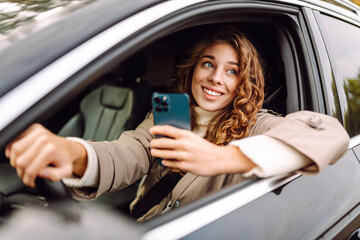A young woman with curly hair enjoys driving by smiling and using her smartphone, capturing a moment of joy on a sunny day, showcasing casual confidence. Leisure, travel, technology. Carsharing.
