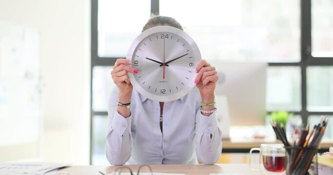 Female employee playfully holds wall clock up to face in office. Woman worker cheerful act demonstrates readiness to end day at workplace