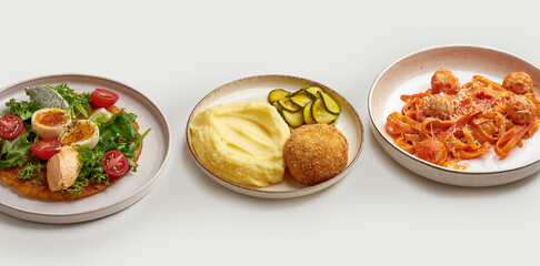 A variety of delicious and appetizing dishes are arranged on a white surface. The dishes include a bowl of creamy soup, a plate of meatballs and pasta, a plate of mashed potatoes