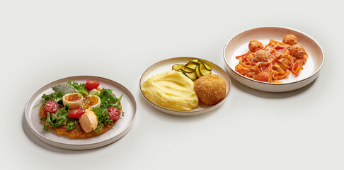 A variety of delicious and appetizing dishes are arranged on a white surface. The dishes include a bowl of creamy soup, a plate of meatballs and pasta, a plate of mashed potatoes