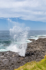 A powerful jet of water erupts from the Little Blowhole in Kiama, New South Wales, Australia, against a backdrop of blue sky and crashing waves.