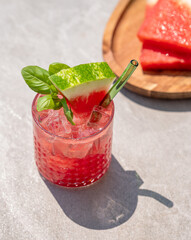 Glass of refreshing watermelon cocktail with basil and ice with fruit slices and fresh herb on grey concrete background with shadow.