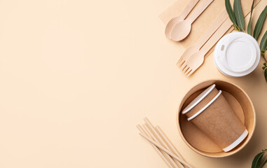 Set of eco-friendly disposable tableware with wooden cutlery, brown paper cups, bowls and bamboo straws on beige background with leaves.