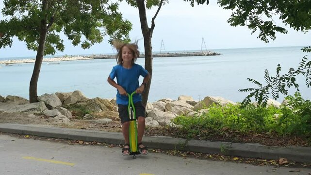 A boy in a blue shirt enjoys hopping on a pogo stick by the seaside, with trees and rocks in the background. The calm sea and distant pier add a peaceful vibe