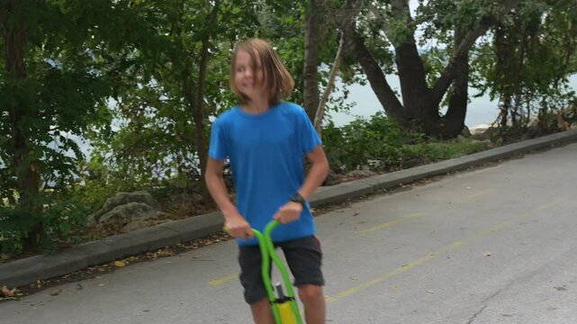 A young boy on a green pogo stick plays joyfully by the seaside, near rocks and trees. The tranquil ocean and a distant pier create a serene and adventurous atmosphere