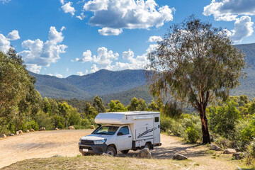 A camper van is parked beneath a large tree at a campsite surrounded by lush greenery and...