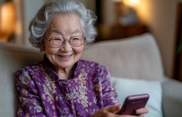 An older woman with silver hair and glasses, dressed in purple, sits on a sofa smiling in front of her phone