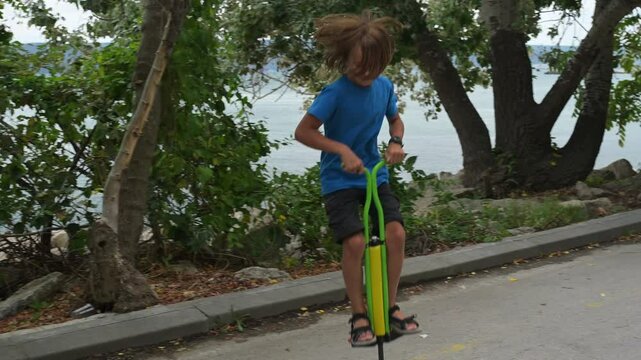 A young boy on a green pogo stick plays joyfully by the seaside, near rocks and trees. The tranquil ocean and a distant pier create a serene and adventurous atmosphere