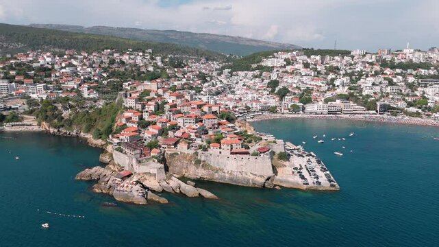 Flying close to the fortress of Ulcinj with the busy touristy beach and bay with its white houses, landscape, cityscape and blue adriatic sea water in the background showing the narrow streets