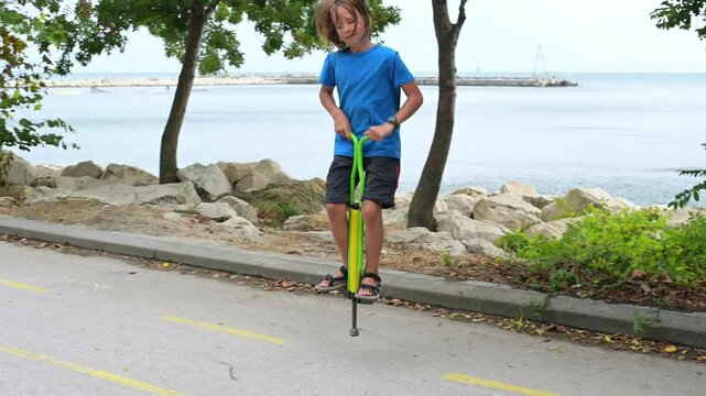 A boy in a blue shirt enjoys hopping on a pogo stick by the seaside, with trees and rocks in the background. The calm sea and distant pier add a peaceful vibe