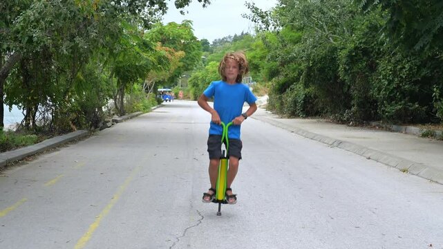 A young boy on a green pogo stick plays joyfully by the seaside, near rocks and trees. The tranquil ocean and a distant pier create a serene and adventurous atmosphere