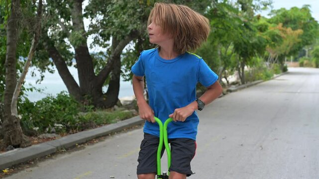 A young boy on a green pogo stick plays joyfully by the seaside, near rocks and trees. The tranquil ocean and a distant pier create a serene and adventurous atmosphere
