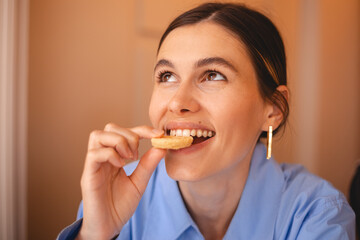 Happy woman eating cookie sitting in a restaurant. Positive woman bites cookie enjoys eating sweet food breaks diet. Woman wear blue shirt, low ponytail rest in cafe. Girl bite cake and look happy.