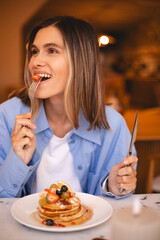 Happy woman eating strawberry with fork, pancakes with fruit and maple syrup in a restaurant interior and looks at side. Close up happy girl with blonde hair look enjoying in cafe. Hold knife, fork.