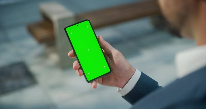 Male Hand Holding a Smartphone with Green Screen Mock Up Display. Man is Standing on a Street and Checking Content on a Mobile Device. Close Up Over the Shoulder Footage