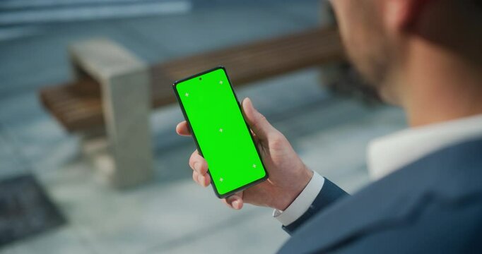 Man Holding a Green Screen Mock Up Chroma Key Smartphone on a Street. Over the Shoulder Footage of a Person Using a Mobile Phone. Template for Internet Content, Social Media, Online Shopping App