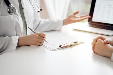 Doctor and patient sitting and discussing something near each other at the white desk in clinic. Female physician is pointing into laptop screen. Medicine concept