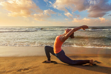 Yoga outdoors - sporty fit woman practices yoga Anjaneyasana - low crescent lunge pose outdoors at beach on sunset