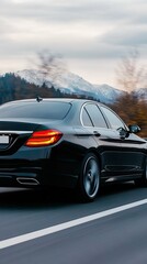 Naklejka premium A black car races along a winding road, framed by majestic snow-covered mountains and lush forests, showcasing speed and motion against a dramatic backdrop