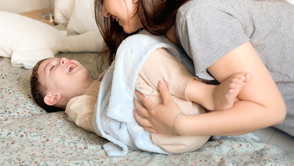 Mom tickles the feet of her little son, wrapped in a blue blanket. Mom and child have fun together in a cozy bedroom at home. Happy children with mother. Bright photos of the baby's childhood