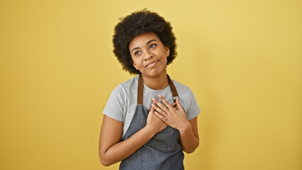 A beautiful adult black woman with curly hair, wearing a denim apron, stands against a yellow background, expressing gratitude with a hand on her chest.