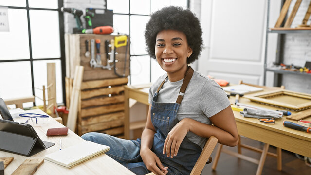 Smiling african american woman in casual attire sitting in a well-equipped carpentry workshop indoors.