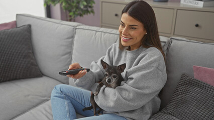 A smiling woman casually reclines on a sofa with a chihuahua in her lap, holding a remote in a modern living room.