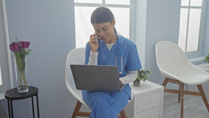 A hispanic nurse in scrubs uses a smartphone and laptop in a bright hospital room.