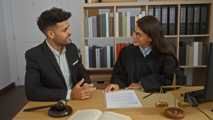 Man and woman in office discussing legal documents with scales of justice and gavel on desk indicating a professional legal environment like a law firm or court