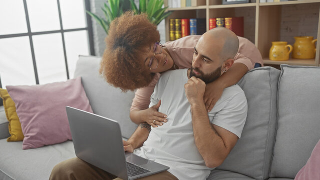 Interracial couple relaxing together on a gray sofa with a laptop in a cozy living room interior