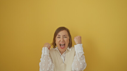 Excited mature woman with short hair celebrates, clenching fists against a vibrant yellow...