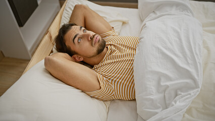 A thoughtful young hispanic man with a beard lies in bed, gazing upward in a cozy bedroom setting.