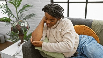 Young african american woman with dreadlocks feeling elbow pain while sitting on a sofa indoors