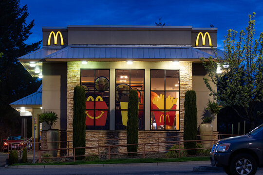Beaverton, OR, USA - April 2, 2024 -  McDonald's drive thru restaurant with its signs and symbols, next to the American flag waving in wind, captured at stormy twilight. High quality picture download
