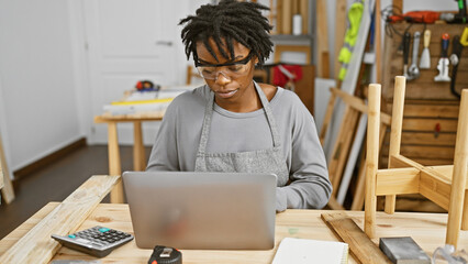 Focused african american woman with dreadlocks working on a laptop in a woodworking workshop.