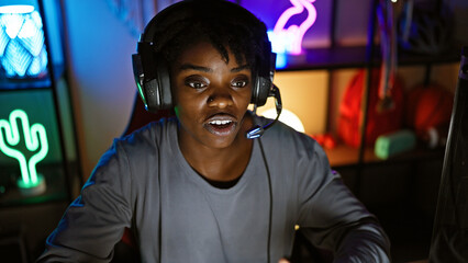 Focused african american woman wearing headphones gaming at night in a colorful neon-lit room.