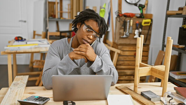 Young black woman with glasses and dreadlocks sits thoughtfully at a wooden desk in a carpentry workshop.