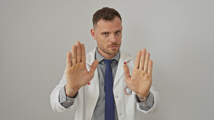 Serious young man in lab coat and tie making stop gesture with both hands, against a white wall...