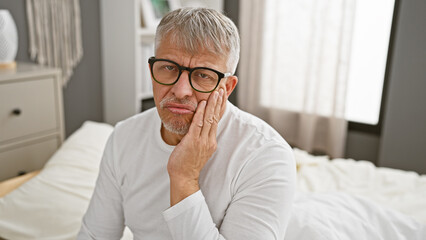 A pensive, grey-haired man wearing glasses sits on a bed in a bedroom, giving a thoughtful expression to the camera.