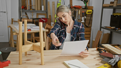 A focused young woman examines paperwork in a cluttered carpentry workshop while on the phone.