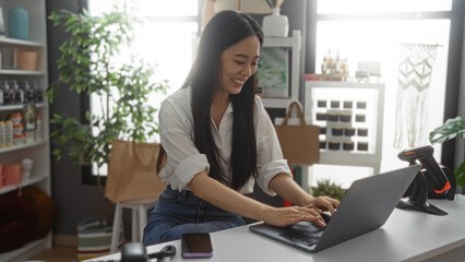 Woman using laptop in home decor shop, smiling at desk surrounded by beautiful decorations and plant, showcasing cheerful work environment.