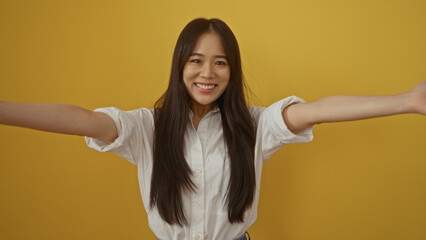 Young chinese woman smiling against yellow background, spreading arms for a welcoming gesture,...
