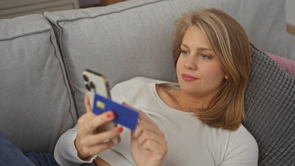 Blonde woman sitting on a couch indoors, holding a smartphone and credit card, contemplating an...